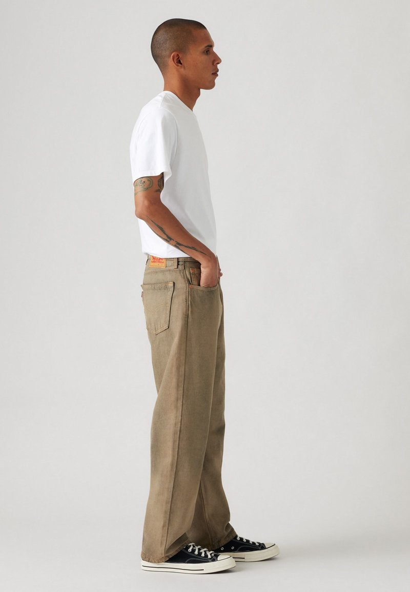 Camiseta blanca ligera combinada con pantalones khaki sueltos que presentan una textura sutil. Zapatillas negras completan el atuendo.