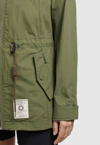 Giacca verde oliva con chiusura a zip, vita regolabile, tasca con bottone e etichetta in tessuto sulla parte anteriore sinistra. Materiale leggero e strutturato.