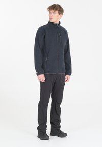 Fleece-takki tummanharmaana, jossa on vetskari edessä, korkea kaulus ja kaksi sivutaskua vetskarilla. Yhdistetty mustiin pantsuihin ja tummiin vaelluskengiin.