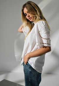 Bluză albă din bumbac cu detalii decorative din dantelă cu ochiuri pe mâneci, asociată cu blugi din denim albastru. Decupajul gulerului este rotund și ușor lejer.