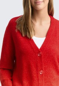 Czerwony dziergany sweter na guziki z dekoltem w serek, posiadający dwa guziki i miękką, teksturowaną tkaninę. Nosi się go na białą bluzkę.