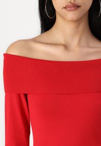 Czerwona bluzka typu off-shoulder z prążkowaną teksturą oraz długimi rękawami, z szerokim prążkowanym dekoltem, który podkreśla ramiona.