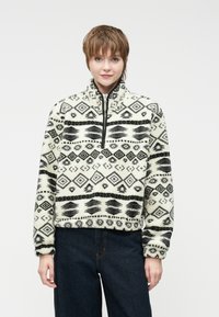 Avių vilnos puloveris kreminės spalvos su juodais geometriniais raštais, aukšta apykakle, pusiau užtrauktuku priekyje ir minkšta, tekstūruota medžiaga. Trumpas kirpimas.