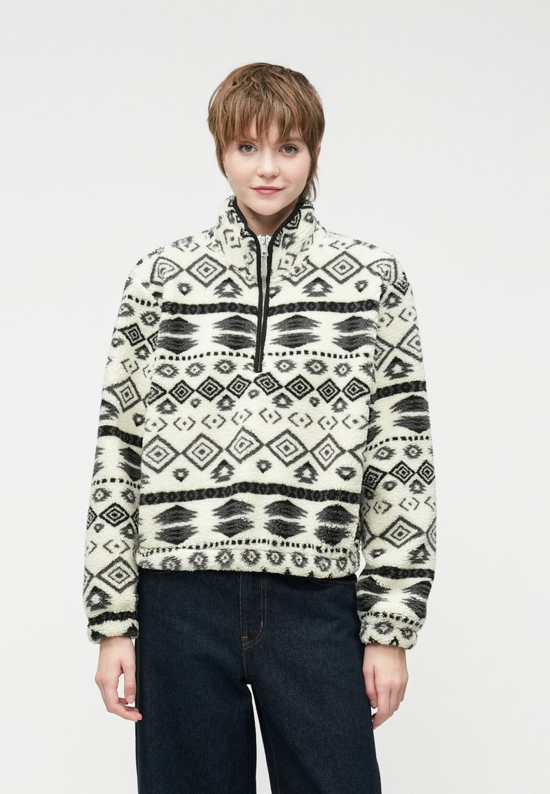 Avių vilnos puloveris kreminės spalvos su juodais geometriniais raštais, aukšta apykakle, pusiau užtrauktuku priekyje ir minkšta, tekstūruota medžiaga. Trumpas kirpimas.