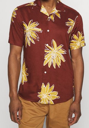 Mężczyzna noszący koszulę na krótki rękaw zapinaną na guziki w ciemnoczerwonym kolorze z dużymi żółtymi kwiatowymi wzorami oraz beżowe spodnie.