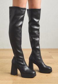 Black thigh-high boots made of smooth leather with a chunky platform and block heel; features a side zipper and seamless upper design.