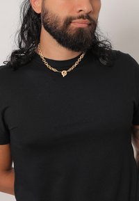 Goldene Kettenhalskette mit ineinandergreifenden Gliedern und einem runden Anhänger, getragen über einem schwarzen T-Shirt mit kurzen Ärmeln und Rundhalsausschnitt.