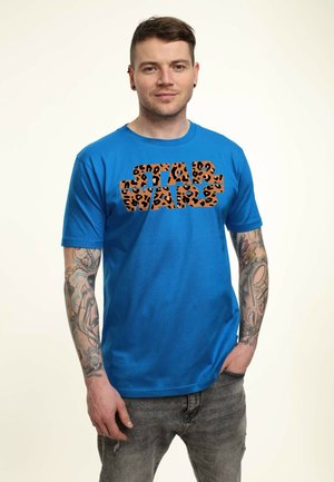 Młody mężczyzna z tatuażami na ramionach stoi, ubrany w niebieską koszulkę z napisem "Star Wars" w panterkę i wyblakłe szare dżinsy, z ręką w kieszeni.