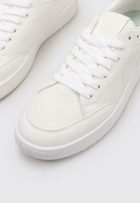 Baskets en toile blanches avec surface texturée, bout rond, lacets plats blancs et un design minimaliste. Semelle en caoutchouc avec un branding subtil.