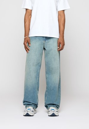 Osoba w jasnoniebieskich szerokich dżinsach, białym t-shircie i białych trampkach z niebieskimi i szarymi akcentami stoi na tle jednolitego tła.