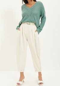 Pull en maille turquoise avec col en V et motif texturé, associé à un pantalon plissé crème à rayures. Sandales blanches à talons ouvertes.