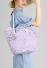 Lavendel Spitzen-Tote-Bag mit komplexem Paisleymuster, silberner Hardware und doppelten Schultergurten; strukturiertes, offenes Design mit dekorativem Anhänger.