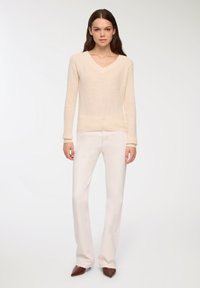 Kremowy sweter dziany z dekoltem w serek, długimi rękawami i teksturowym wykończeniem, w połączeniu z jasnymi spodniami typu dzwony i brązowymi, spiczastymi butami.
