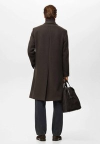 Abrigo largo de lana marrón con cuello alto, abertura única y silueta entallada. Combinado con jeans oscuros y un bolso de ante negro.