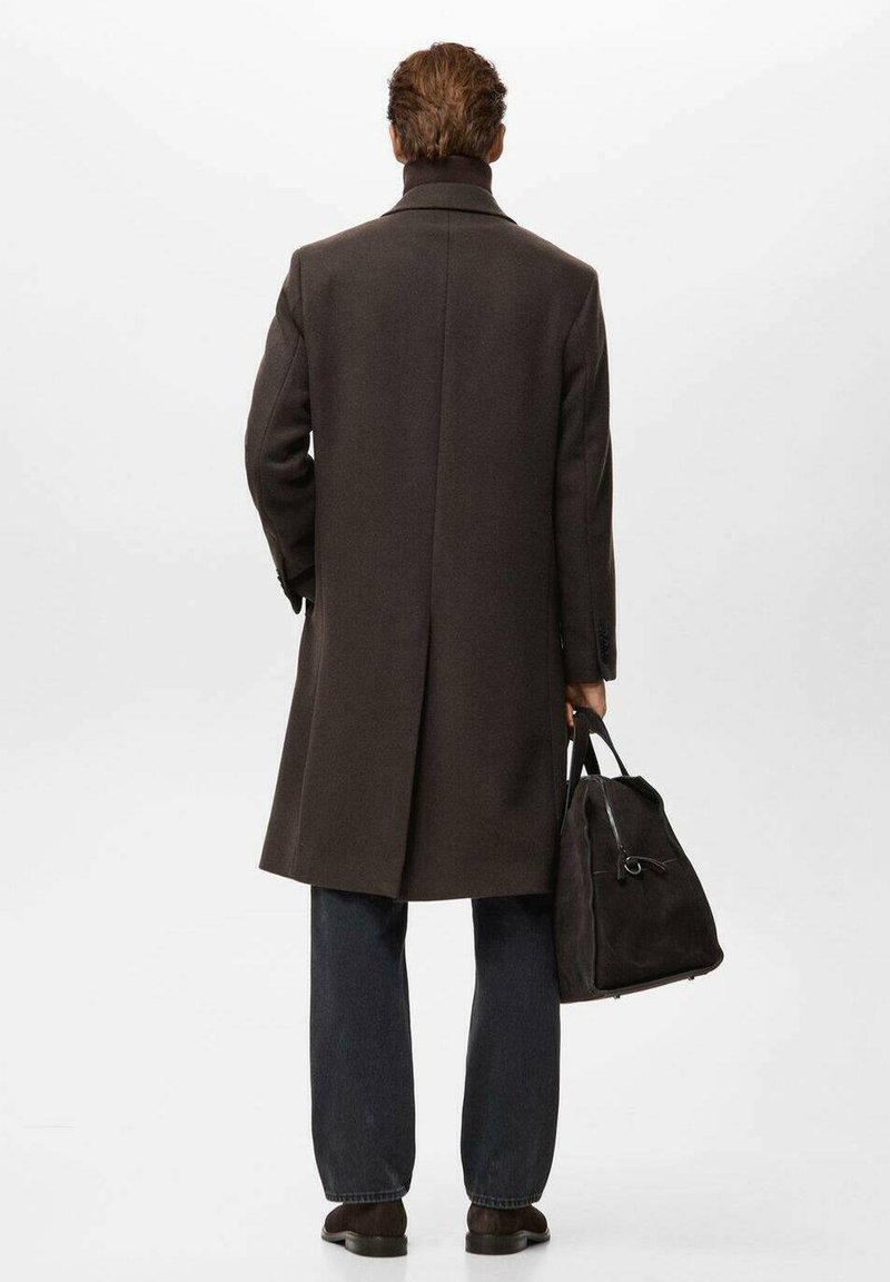 Lungo cappotto di lana marrone con collo alto, spacco singolo e silhouette sagomata. Abbinato a jeans scuri e una borsa nera in camoscio.