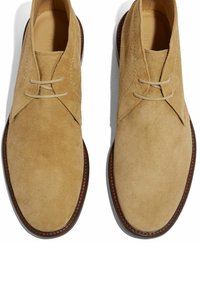 Bež nubuk čizme chukka s dva vezna otvora, zaobljenim prstom i kontrastnom smeđom kožnom potplatom. Glatka unutrašnja podstava.