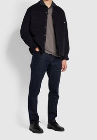 Veste en velours côtelé foncé avec deux poches poitrine sur une chemise en mesh texturé, associée à un jean foncé et des chaussures noires à lacets.