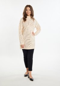 Beige Strick-Tunika-Pullover mit wellenförmigen silbernen Akzenten, kombiniert mit schwarzen schmalen Hosen und schwarzen Absatzschuhen. Glatte Textur, hoher Kragen.