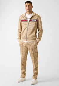 Beige trainingspak met bijpassend vest met rits en broek, voorzien van geperste logopatronen, rode en zwarte streepaccenten en zijzakken.