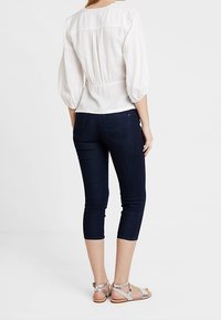 Mujer vistiendo unos jeans skinny recortados de color azul oscuro, blusa blanca de manga tres cuartos y sandalias con tiras plateadas, de pie dando la espalda.