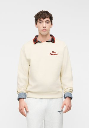 Noor mees, kes kannab kreemjat New Balance'i pusad punase ja musta ruudulise särgi ja helehallide New Balance'i pükstega, seisab ühevärvse tausta ees.