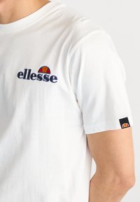 Camiseta blanca de manga corta hecha de algodón, cuenta con un logo bordado de "ellesse" en azul marino y rojo, con una pequeña etiqueta del logo en la manga.