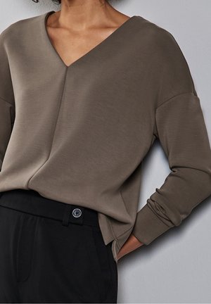 Person trägt ein lockeres, langärmeliges braunes V-Ausschnitt-Oberteil, das in hoch taillierte schwarze Hosen mit einem Knopf am Bund gesteckt ist, Hand in der Tasche.