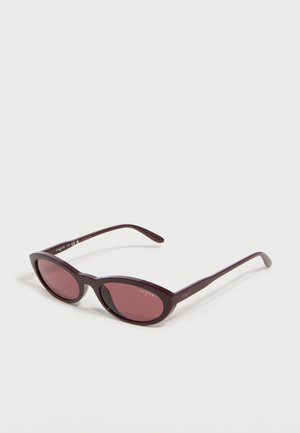 Gafas de sol estilo ojo de gato con montura morado oscuro y lentes tintadas, con la marca "Vogue" visible en las varillas y en la lente.