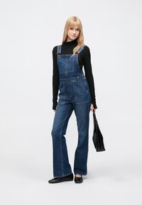 Jeans kombinezon z ohlapnim krojem, temno modre barve, nastavljivi trakovi, sprednji žepi, kombiniran s črnim dolgimi rokavi in črnimi dodatki.