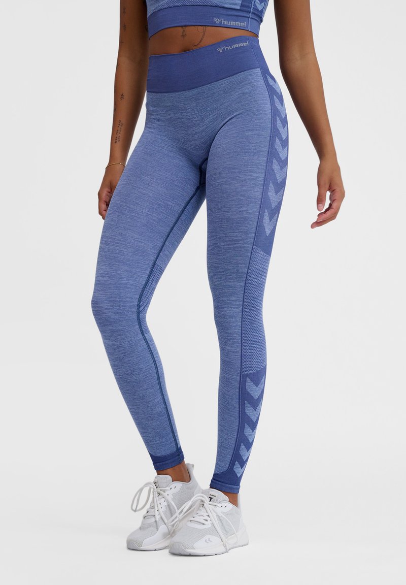 Leggings sportivi blu con vita alta, tessuto testurizzato e motivo a chevron lungo i lati. Indossati con scarpe da ginnastica bianche.