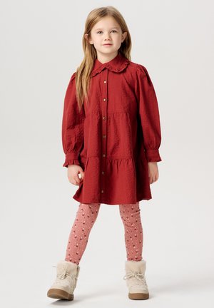 Rotes Hemdkleid mit rüschengem Kragen, strukturiertem Stoff und schwingender Silhouette, kombiniert mit pinken gemusterten Leggings und beigen Stiefeln.