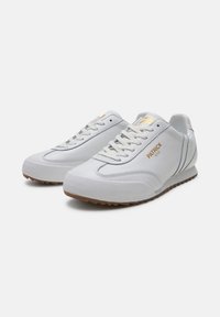 Weiße Sport Schuhe aus strukturiertem Leder und synthetischen Akzenten, ausgestattet mit einer Gummisohle, Schnürverschluss und goldfarbenem Logodetail an der Seite.