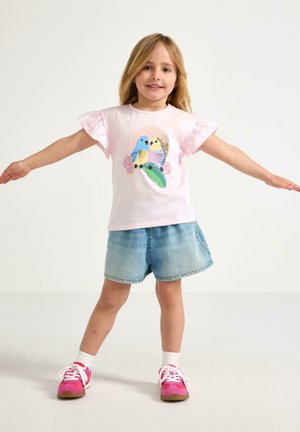 Ung pige står med armene udstrakte iført lyserød T-shirt med farverigt fugledesign, denimshorts, hvide sokker og lyserøde sneakers.
