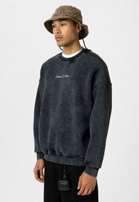 Donkergrijs, oversized sweatshirt met geborduurde tekst "De Toekomst Is Hier" op de borst, gecombineerd met een tan kleuren lederen bucket hat en zwarte broek.