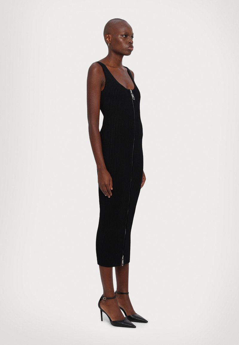 Robe midi noire côtelée avec une fermeture éclair devant, dotée de fines bretelles. Associée à des talons aiguilles noirs avec des brides au niveau de la cheville.