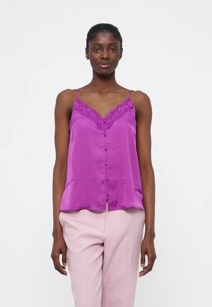 Kvinne som har på seg en lilla spilekantet camisole med knapper foran og lyserosa bukser, stående mot en enkel hvit bakgrunn.