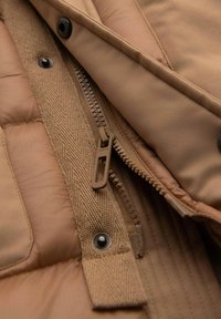 Tan pufferjack interior met gestikte textuur, zichtbare rits en stofaccenten. Voorzien van zwarte drukknoopjes en een gladde voering.