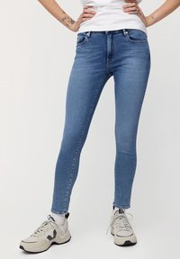 Vaqueros ajustados de mezclilla azul con cinco bolsillos, presillas para el cinturón y una sutil textura descolorida. Combinados con zapatillas deportivas en tonos neutros.