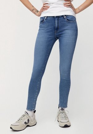 Vaqueros ajustados de mezclilla azul con cinco bolsillos, presillas para el cinturón y una sutil textura descolorida. Combinados con zapatillas deportivas en tonos neutros.