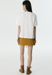 T-shirt en coton blanc à manches courtes associé à une jupe mini jaune moutarde avec des cordons de serrage. Des bottines brunes à la cheville complètent la tenue.