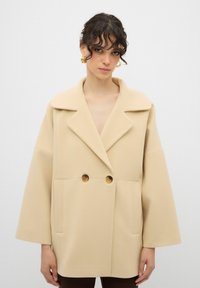 Cappotto oversize beige con colletto a revers, chiusura doppiopetto, grandi bottoni e tasche laterali. Tessuto morbido e testurizzato.