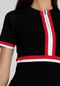 Haut noir à manches courtes avec des rayures rouges et blanches sur les manches, à la taille, et un détail de fermeture éclair vertical au centre devant.