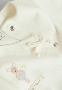 Babykleidung in Cremefarbe mit weichem Stoff, Polka-Dot-Muster, gesticktem Hasendesign, Druckknöpfen und dekorativer Fliege.