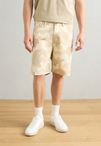 Beige camouflage shorts featuring a relaxed fit, smooth fabric, and back pockets, paired with white sneakers and crew-length socks.