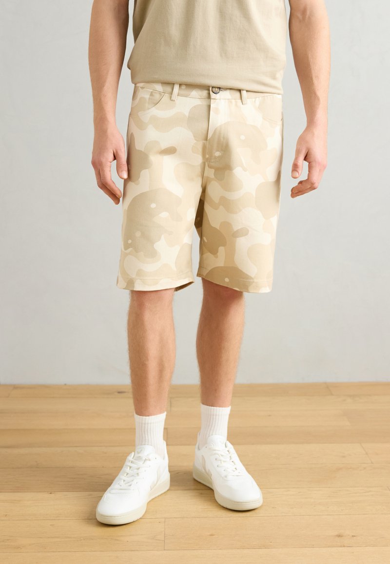 Beige camouflage shorts featuring a relaxed fit, smooth fabric, and back pockets, paired with white sneakers and crew-length socks.