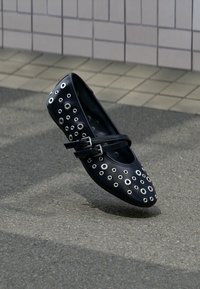 Μαύρη δερμάτινη μπαλαρίνα με ασημένιες οπές, στρογγυλερή μύτη, διακοσμητικό λουράκι με αγκράφα και λεία υφή. Τοποθετημένη σε γκρι επιφάνεια.