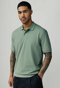 Koszulka polo w przygaszonej zieleni, krótki rękaw, teksturowana tkanina, klasyczny kołnierzyk, zapięcie na dwa guziki, noszona z granatowymi spodniami, ukazująca tatuaże na ramionach.