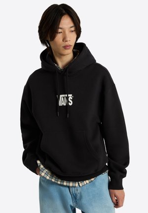 Jeune personne aux cheveux mi-longs foncés portant un sweat à capuche Vans noir par-dessus une chemise à carreaux et un jean bleu clair, regardant vers le bas sur le côté.