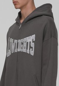 Sudadera con capucha gris y cremallera con "LOWLIGHTS" impreso en blanco. Presenta bolsillos tipo canguro y una tela suave y texturizada.