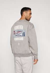 Γκρι φούτερ με άνετη εφαρμογή, διαθέτει γραφικό σχέδιο στην πλάτη με τη φράση «FOREVER FORWARD» και λογότυπο USMNT σε μπλε και κόκκινες αποχρώσεις.